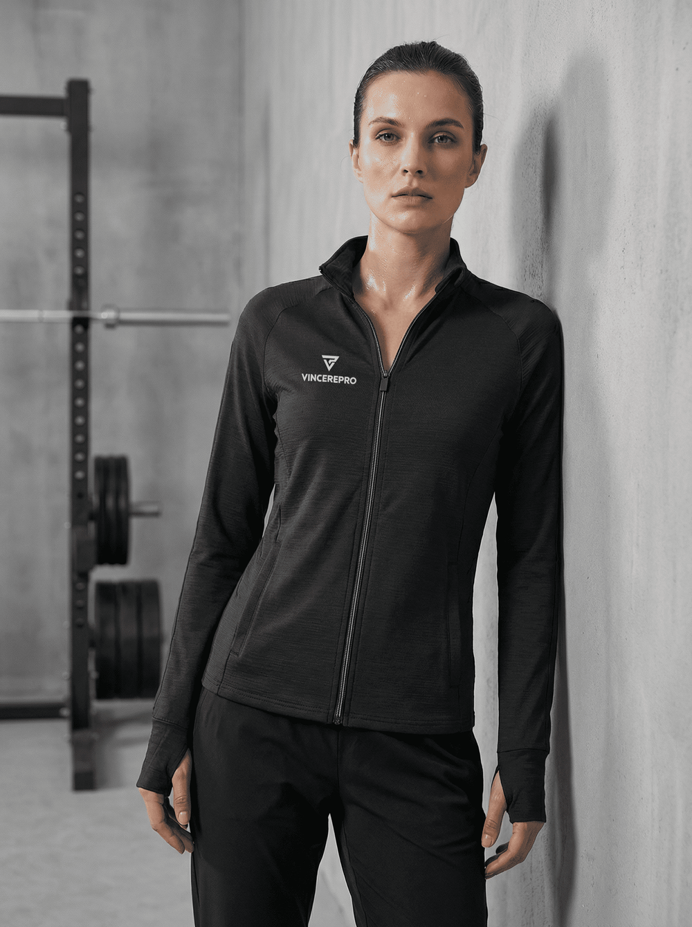 Ženska v črnem športnem puloverju z zadrgo VincerePro stoji ob betonski steni v sodobnem fitnesu, z utežmi in squat rackom v zamegljenem ozadju, minimalističen gym ambient.