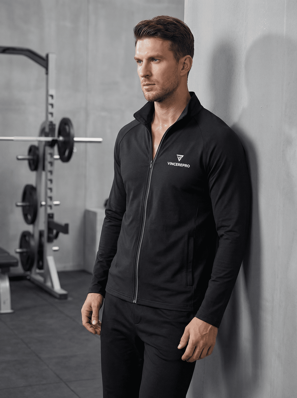 Moški v črnem športnem puloverju z zadrgo VincerePro stoji ob betonski steni v sodobnem fitnesu, z utežmi in squat rackom v zamegljenem ozadju, minimalističen gym ambient.