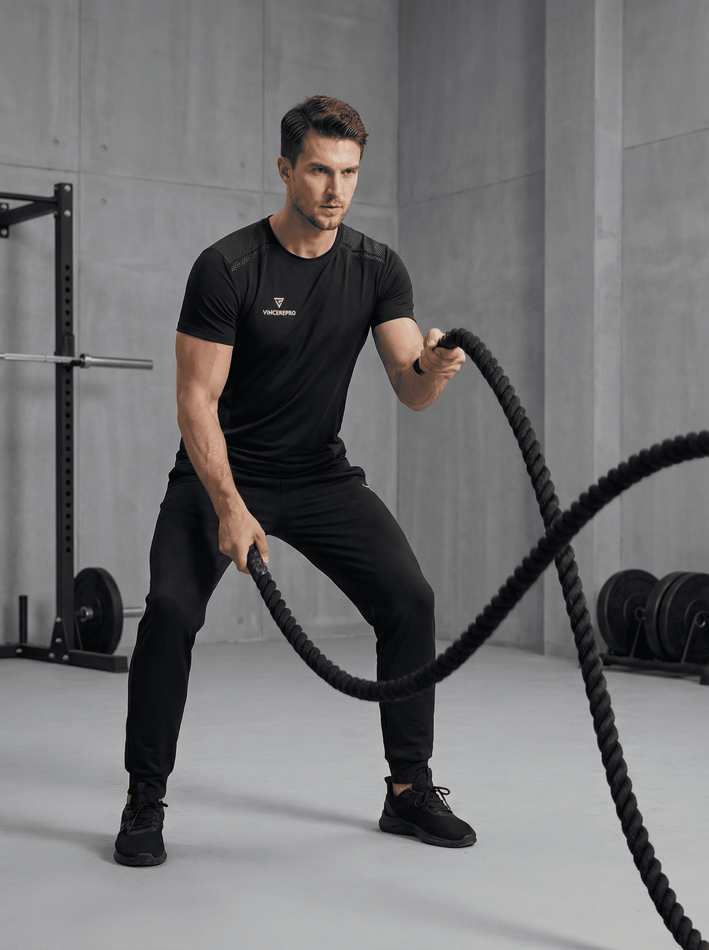 Moški v črni športni majici VincerePro izvaja vajo z bojnimi vrvmi v sodobnem minimalističnem fitnesu, z utežmi in squat rackom v zamegljenem ozadju.
