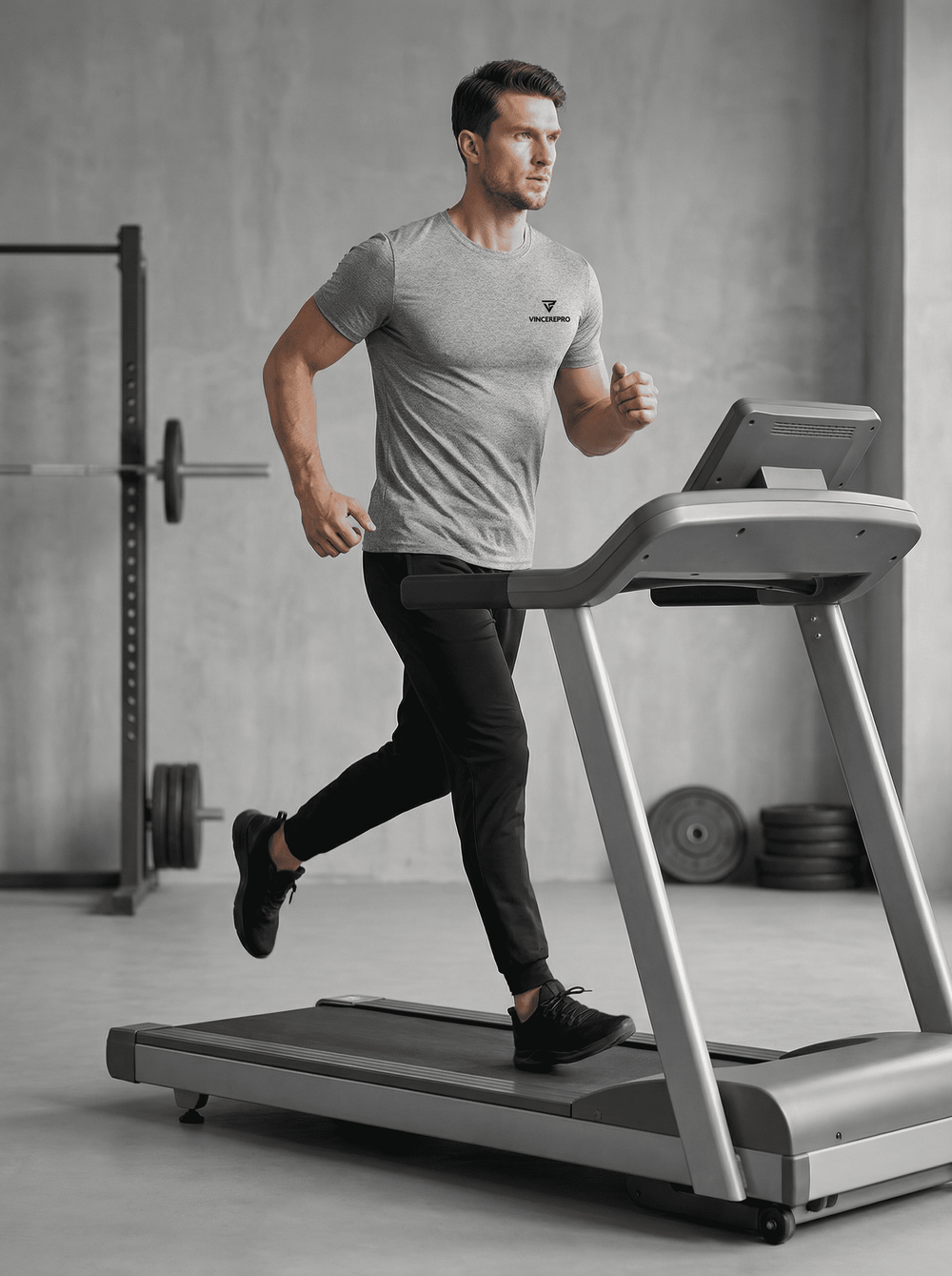 Moški v sivi športni majici VincerePro teče na tekaški stezi v sodobnem minimalističnem fitnesu, z utežmi in squat rackom v zamegljenem ozadju.