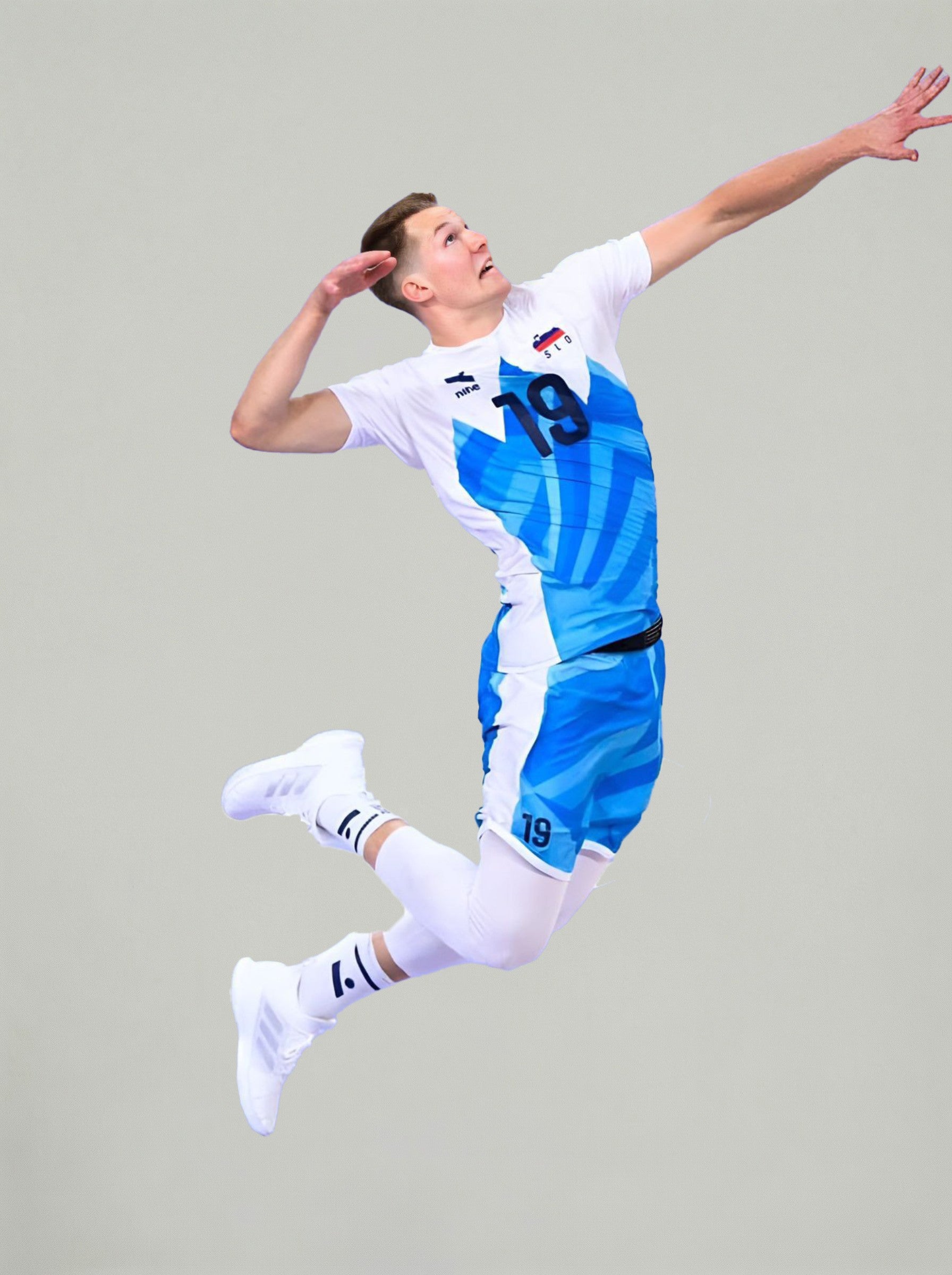 Športnik v modro-belem dresu s številko 19 je v zraku med odbojkarskim skokom, pripravljen na napad ali servis, na nevtralnem svetlem ozadju.