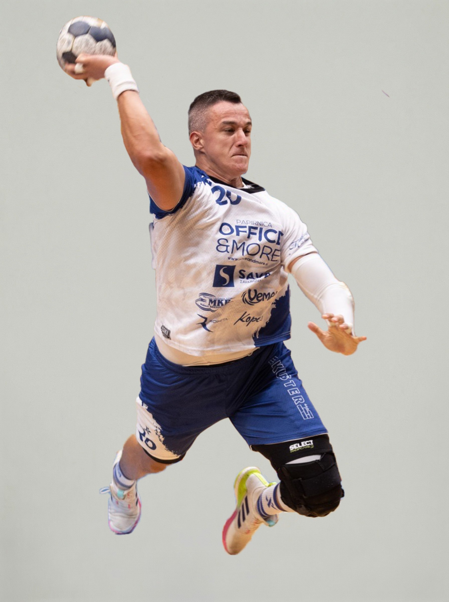 Rokometaš v modro-belem dresu je v zraku med metom na gol, z žogo v dvignjeni roki, na nevtralnem svetlem ozadju.