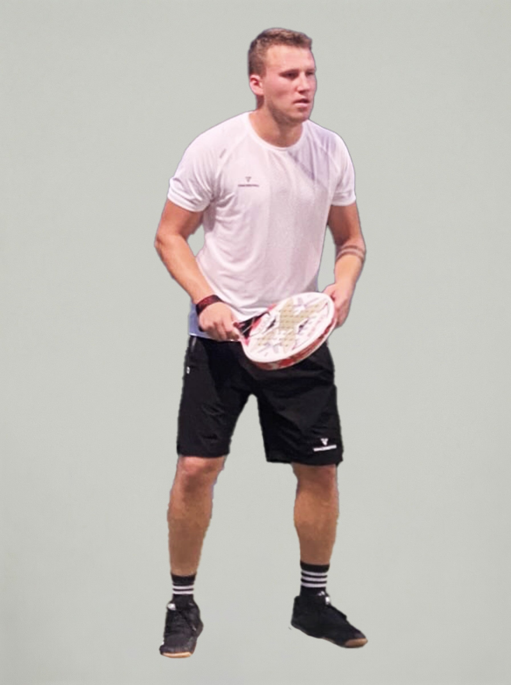Moški v beli športni majici in črnih kratkih hlačah stoji v pripravljenem položaju z loparjem za padel, na nevtralnem svetlem ozadju.