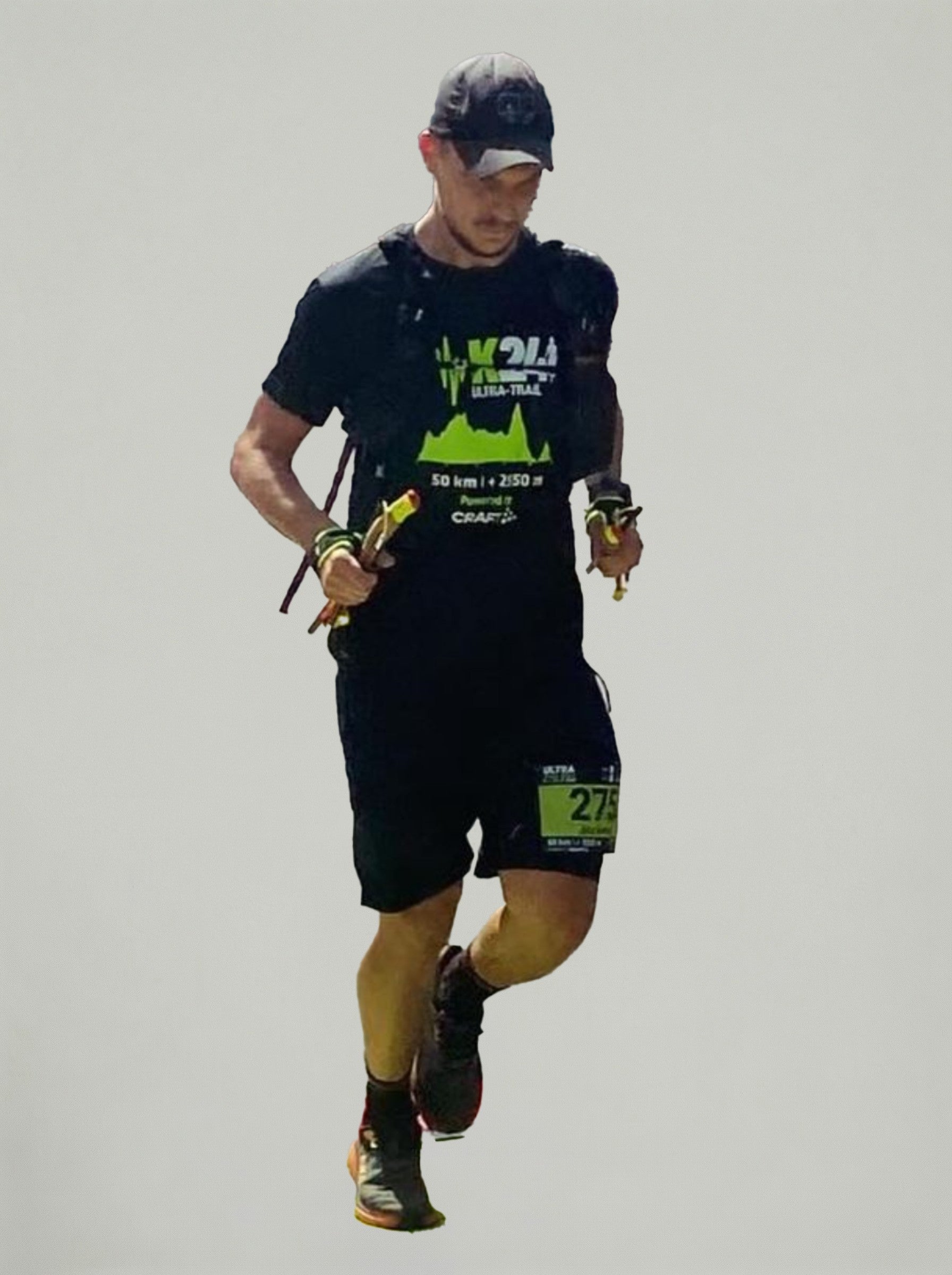 Tekač na trail tekmi v črni športni opravi in kapi teče z nahrbtnikom in tekaškimi palicami, na nevtralnem svetlem ozadju.