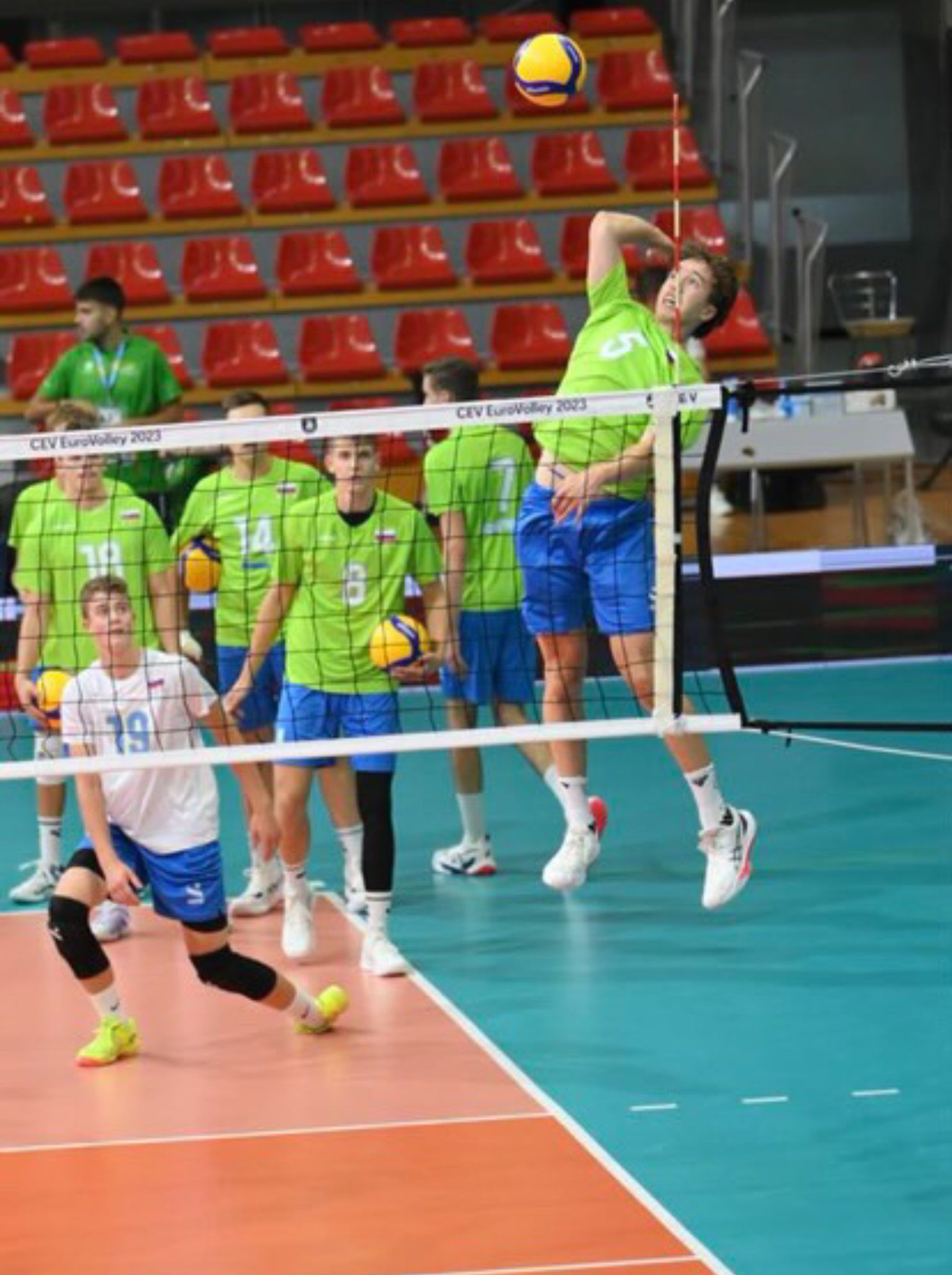 Odbojkar v zelenem dresu skače ob mreži in izvaja napadalni udarec med tekmo v športni dvorani, soigralci in tribune z rdečimi sedeži v ozadju.