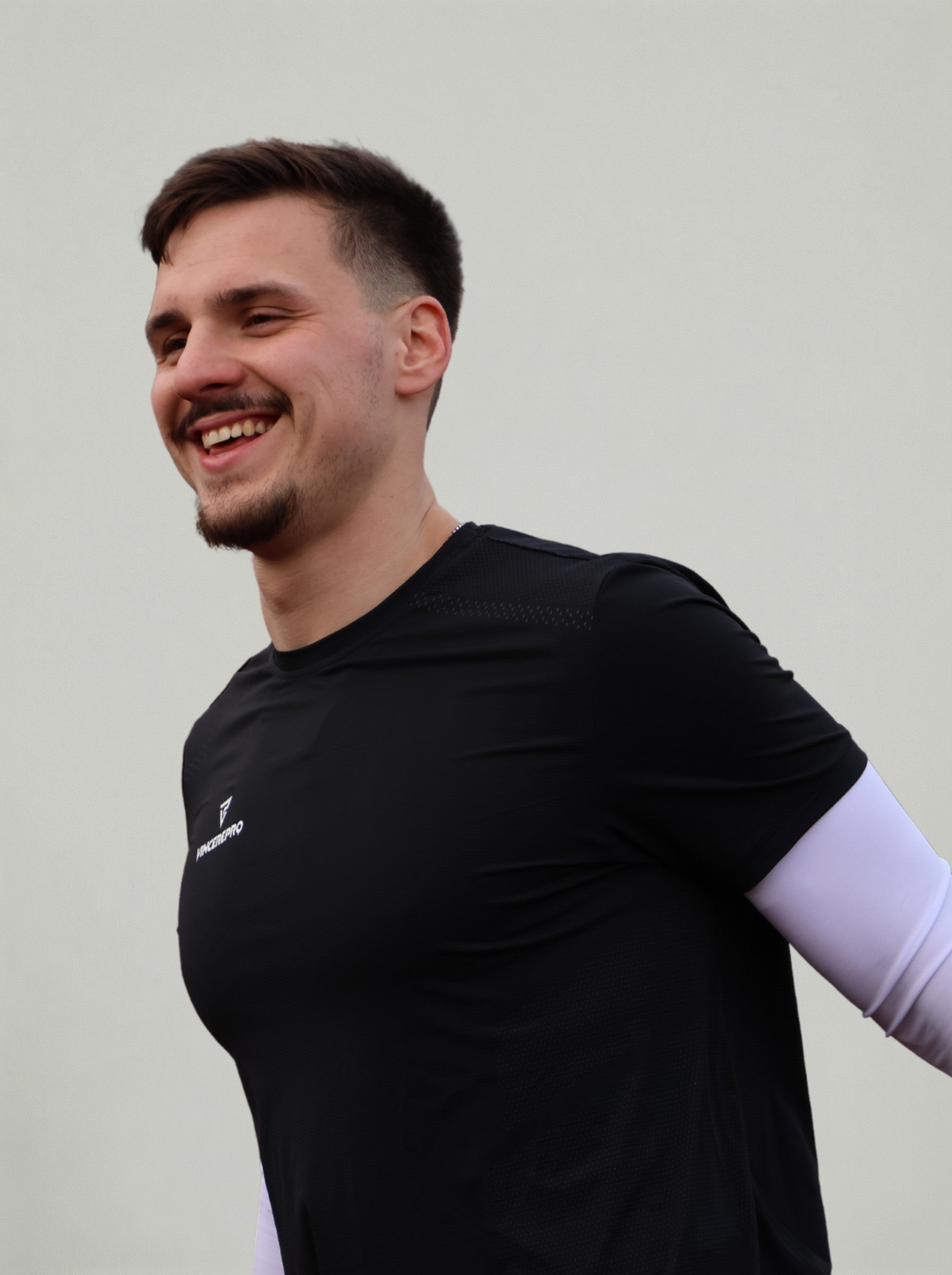 Nasmejan moški v črni športni majici stoji na nevtralnem svetlem ozadju, oblečen v oprijeto funkcionalno športno opremo.