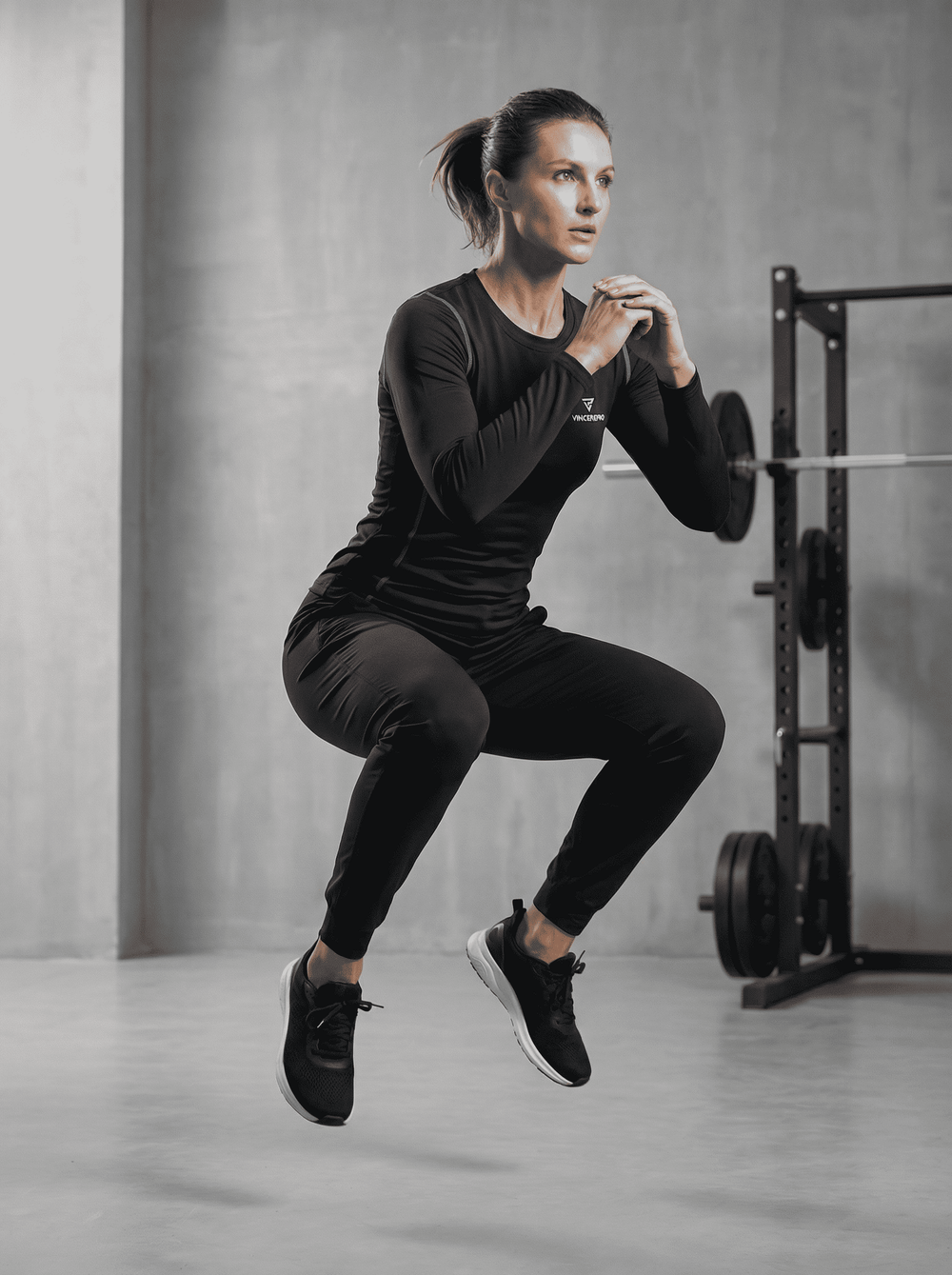 Ženska v črni športni opravi VincerePro izvaja eksploziven počepni skok v sodobnem minimalističnem fitnesu, z utežmi in squat rackom v zamegljenem ozadju.